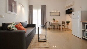 Ein modernes Wohnzimmer mit einem grauen Sofa, einem Essbereich mit weißem Tisch und Stühlen sowie einer Küche mit einem weißen Kühlschrank und einer Klimaanlage.