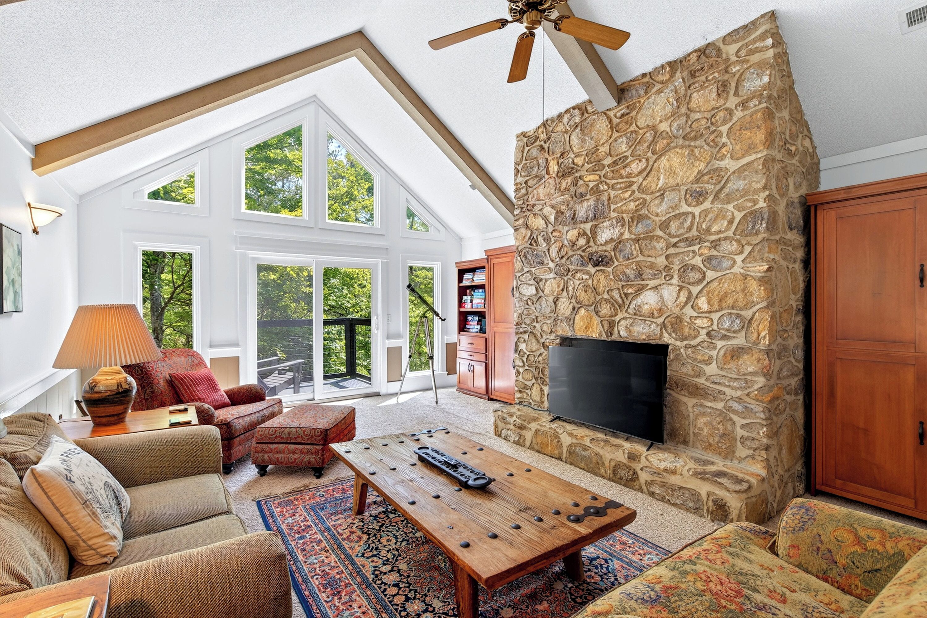 A spacious living room with a stone fireplace, a flat-screen TV, and a large window overlooking a deck.