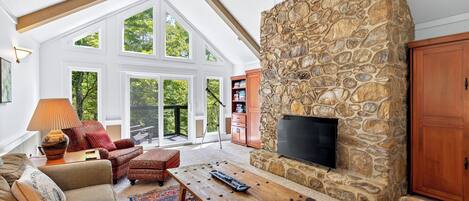 A spacious living room with a stone fireplace, a flat-screen TV, and a large window overlooking a deck.