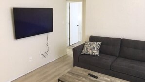 Una sala de estar con un sofá oscuro, una mesa de centro y un televisor de pantalla plana montado en la pared.