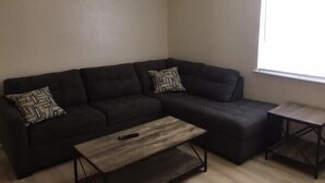 Una sala de estar con un sofá oscuro, dos almohadones estampados, una mesa de centro de madera y una mesita auxiliar.