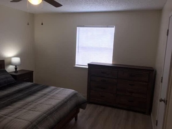 Une chambre à coucher comprenant un lit, une commode, une table de chevet, une fenêtre et un ventilateur de plafond.
