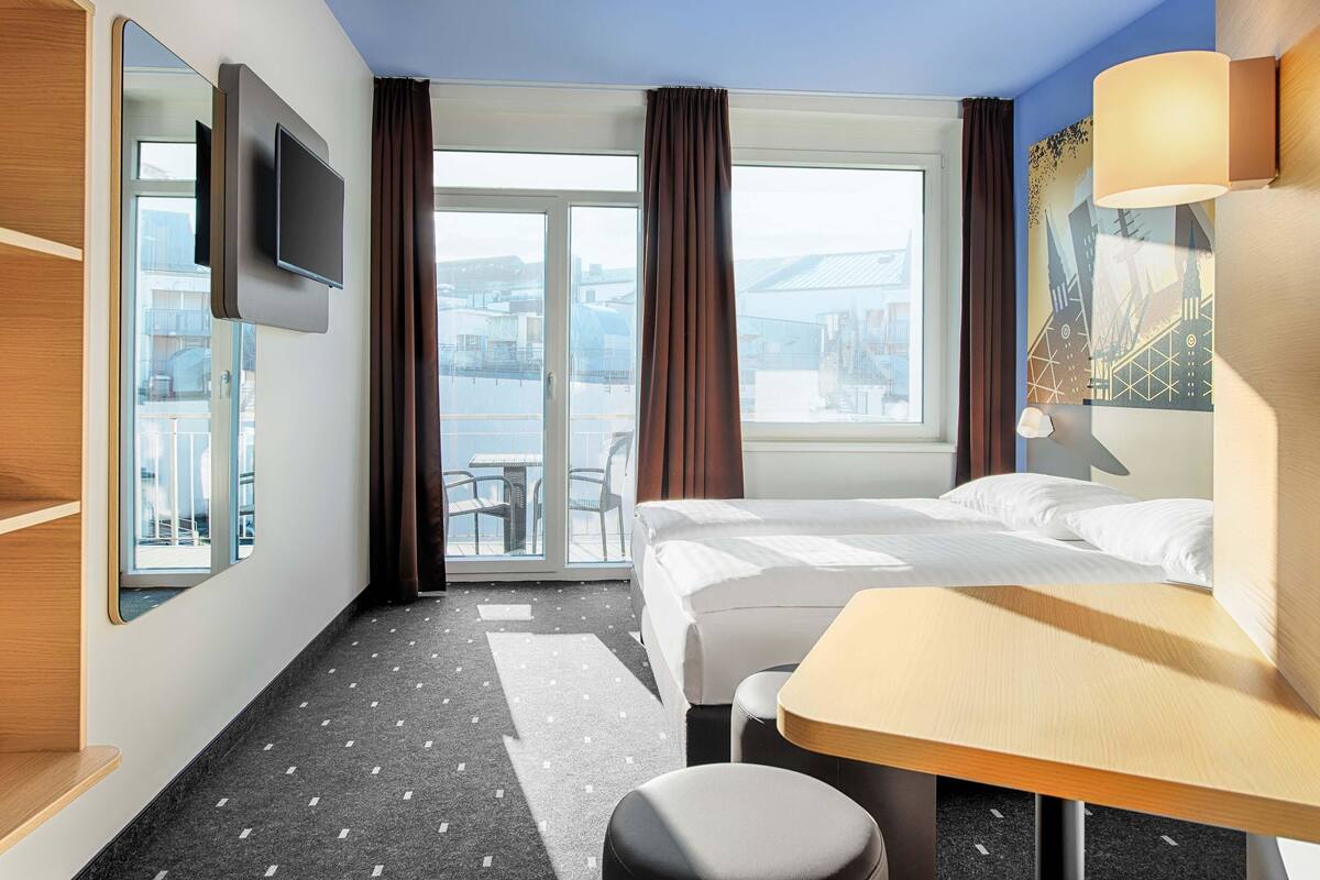 Een hotelkamer met een bed, een bureau, een stoel, een televisie en uitzicht op de stad door grote ramen.
