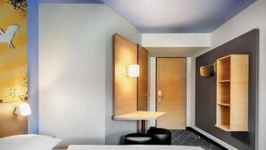 A modern hotel room with a large bed, a wooden wardrobe, a small desk, and a wall-mounted shelf.