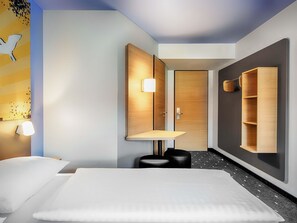 Ein modernes Hotelzimmer mit einem großen Bett, einem Holzschrank, einem kleinen Schreibtisch und einem an der Wand befestigten Regal.