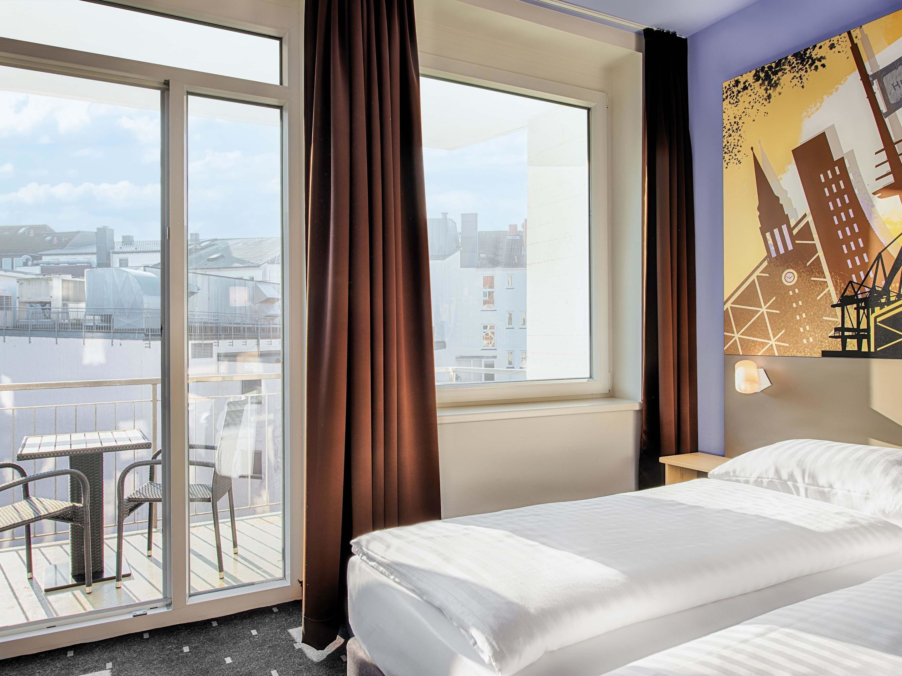 Ein Hotelzimmer mit Bett, einem Vorhangfenster, einem Balkon mit Stühlen und Blick auf die Stadt.