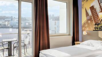 Ein Hotelzimmer mit Bett, einem Vorhangfenster, einem Balkon mit Stühlen und Blick auf die Stadt.