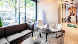 A modern lobby with a sofa, two chairs, a small table with a vase, large windows, and a reception desk.