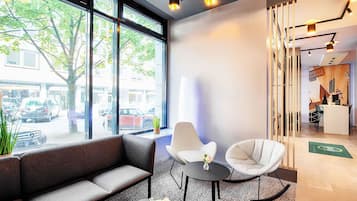 Eine moderne Lobby mit einer Couch, zwei Sesseln, einem kleinen Tisch mit einer Vase, großen Fenstern und einer Rezeption.