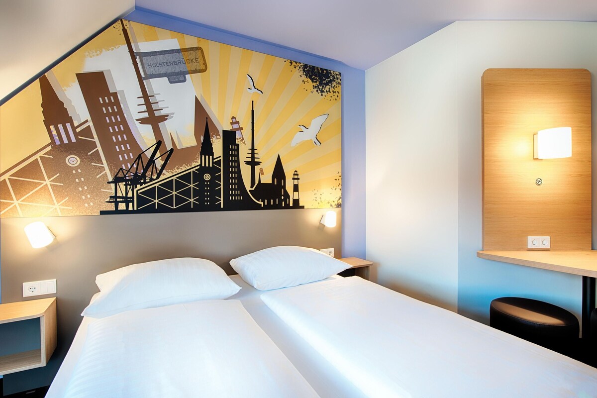 Une chambre d’hôtel avec un lit, une table de chevet, une lampe fixée au mur et une fresque représentant un paysage urbain.