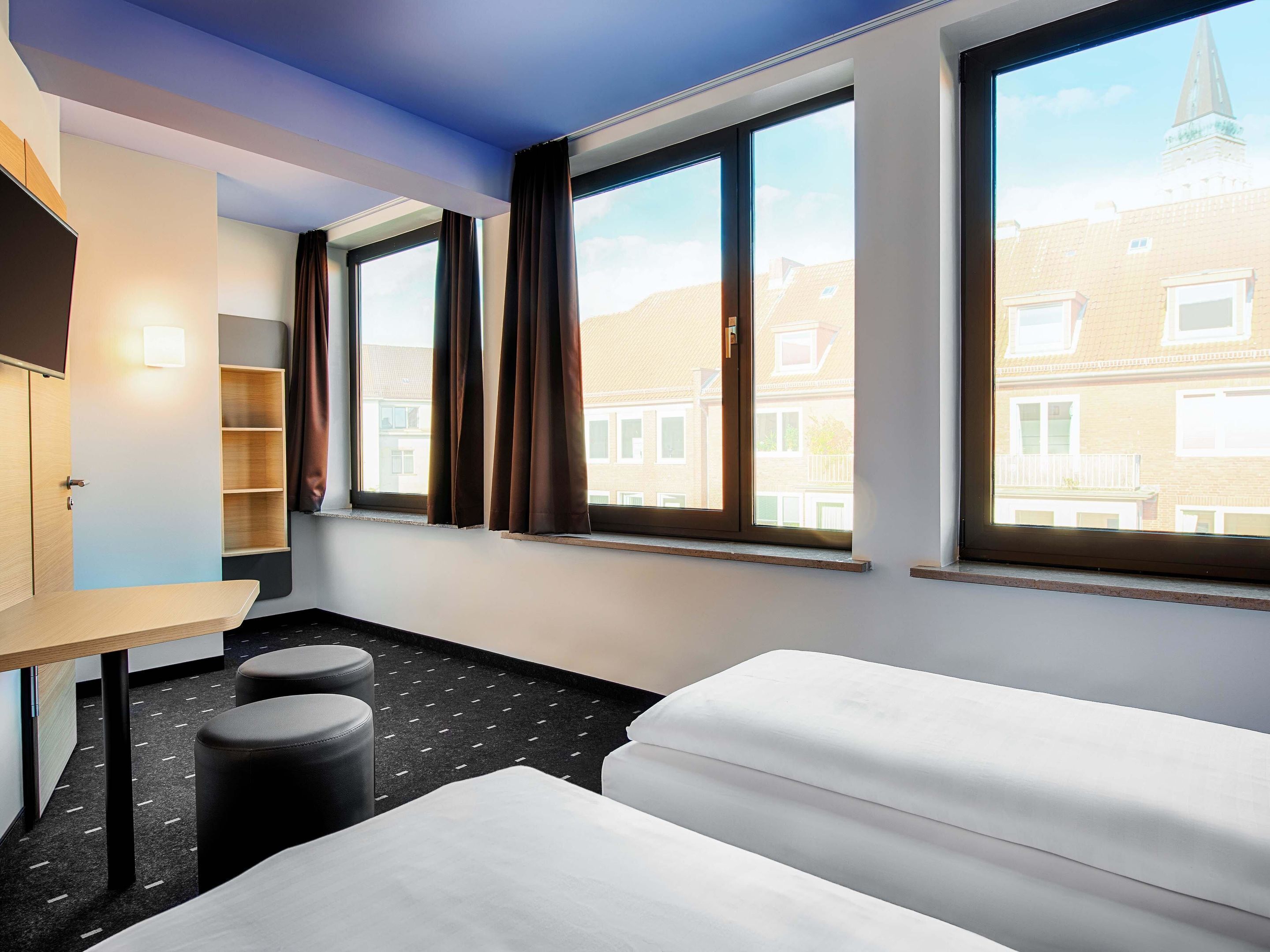 Ein Hotelzimmer mit einem Bett, einem Schreibtisch und einem Stuhl. Es gibt Fenster mit Vorhängen und einen Blick auf Gebäude außerhalb.