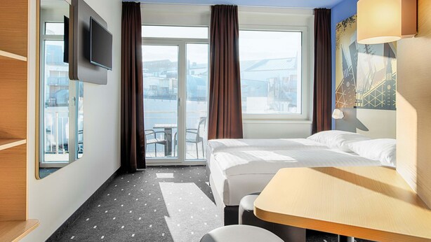 A hotel room with a bed, a desk, a chair, a TV, and a view of the city through large windows.