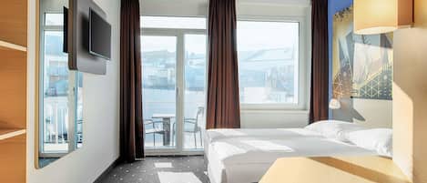 Habitación de hotel con cama, escritorio, silla, televisor y vista de la ciudad a través de amplias ventanas.