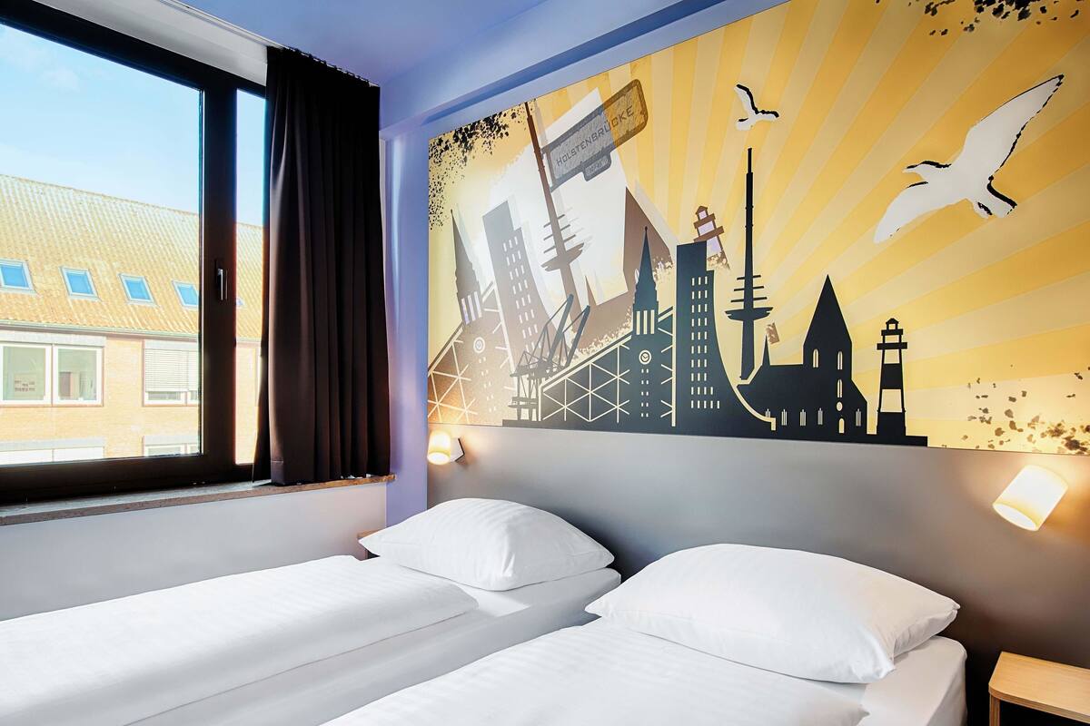 Een hotelkamer met twee bedden, een groot raam en een stadsgezicht muurschildering.