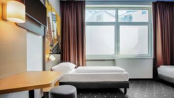Ein Hotelzimmer mit einem Bett, einem Schreibtisch, einem Stuhl, einem kleinen Hocker, einer Wandlampe und einem Fenster mit Vorhängen.
