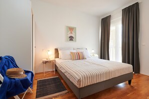 Une chambre à coucher avec un lit, deux tables de chevet, une lampe, un chapeau posé sur une chaise et une fenêtre avec des rideaux.