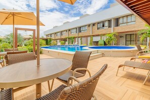 An outdoor pool area with lounge chairs and a table, surrounded by a modern building with large windows and a covered patio. - IT11 Ap 3 Quartos a 50m da Praia (Camaçari)