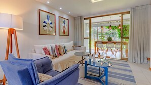 A modern living room with a sofa, a coffee table, and a dining area with chairs. There are framed artworks on the wall and a large window with curtains. - IT11 Ap 3 Quartos a 50m da Praia (Camaçari)