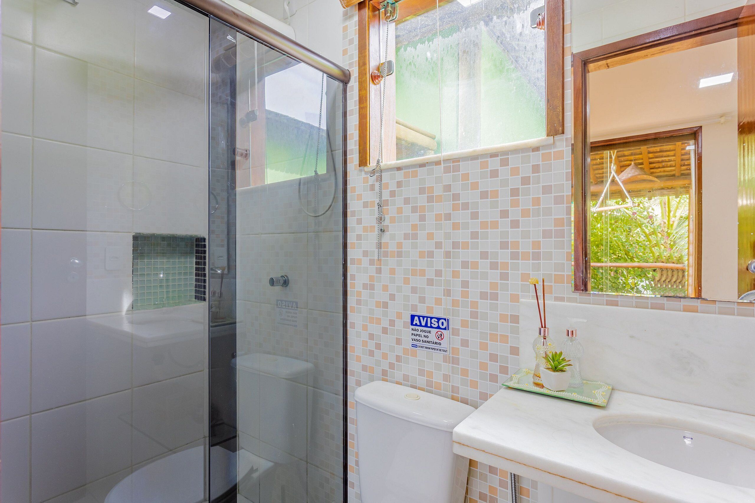 Un baño con cabina de ducha de vidrio, inodoro con salpicadero de azulejos y lavamanos con espejo.