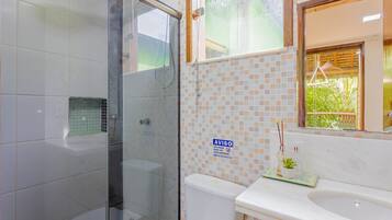 Un baño con cabina de ducha de vidrio, inodoro con salpicadero de azulejos y lavamanos con espejo.