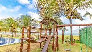 A wooden playground with a slide and swings, surrounded by palm trees and a pool. - IT12 Bangalô Duplex 2 Suítes Pé na Areia (Camaçari)