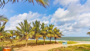 A beach with palm trees, a sandy path, and a small structure. - IT12 Bangalô Duplex 2 Suítes Pé na Areia (Camaçari)