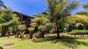 A lush garden with palm trees, a wooden structure, and a green building. - IT12 Bangalô Duplex 2 Suítes Pé na Areia (Camaçari)
