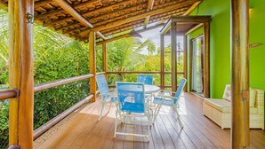 A wooden deck with a table and chairs, a wicker sofa, and a view of lush greenery. - IT12 Bangalô Duplex 2 Suítes Pé na Areia (Camaçari)