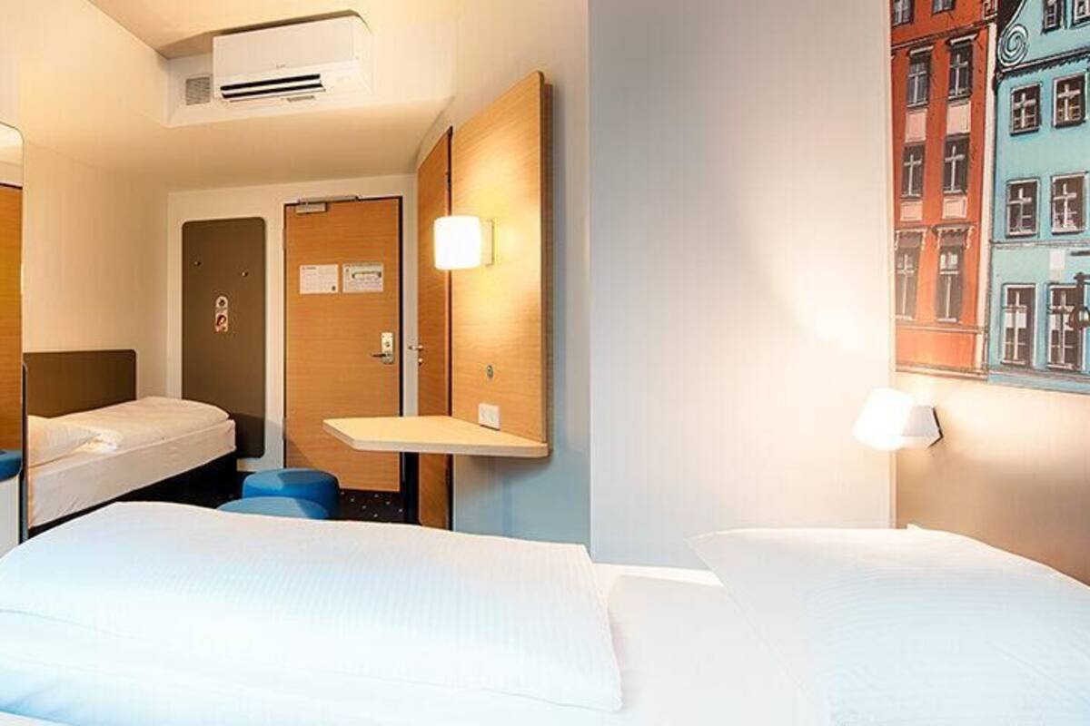 Habitación de hotel con dos camas, un televisor, un espejo, una mesita pequeña y un cuadro en la pared.