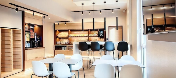 A modern restaurant with white tables and black chairs, a counter with a coffee machine, and a wooden display case with food items.