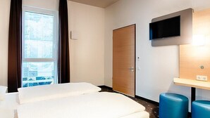 Hotellihuone, jossa on sänky, seinälle asennettu televisio, ikkuna verhoilla ja ovi.