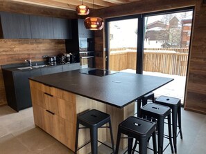 A modern kitchen with a large island, black stools, and a view of a wooden deck outside. - SKI IN SKI OUT PARADISKI LA PLAGNE LES COCHES TOP OF THE RANGE CHALET (Bellentre)