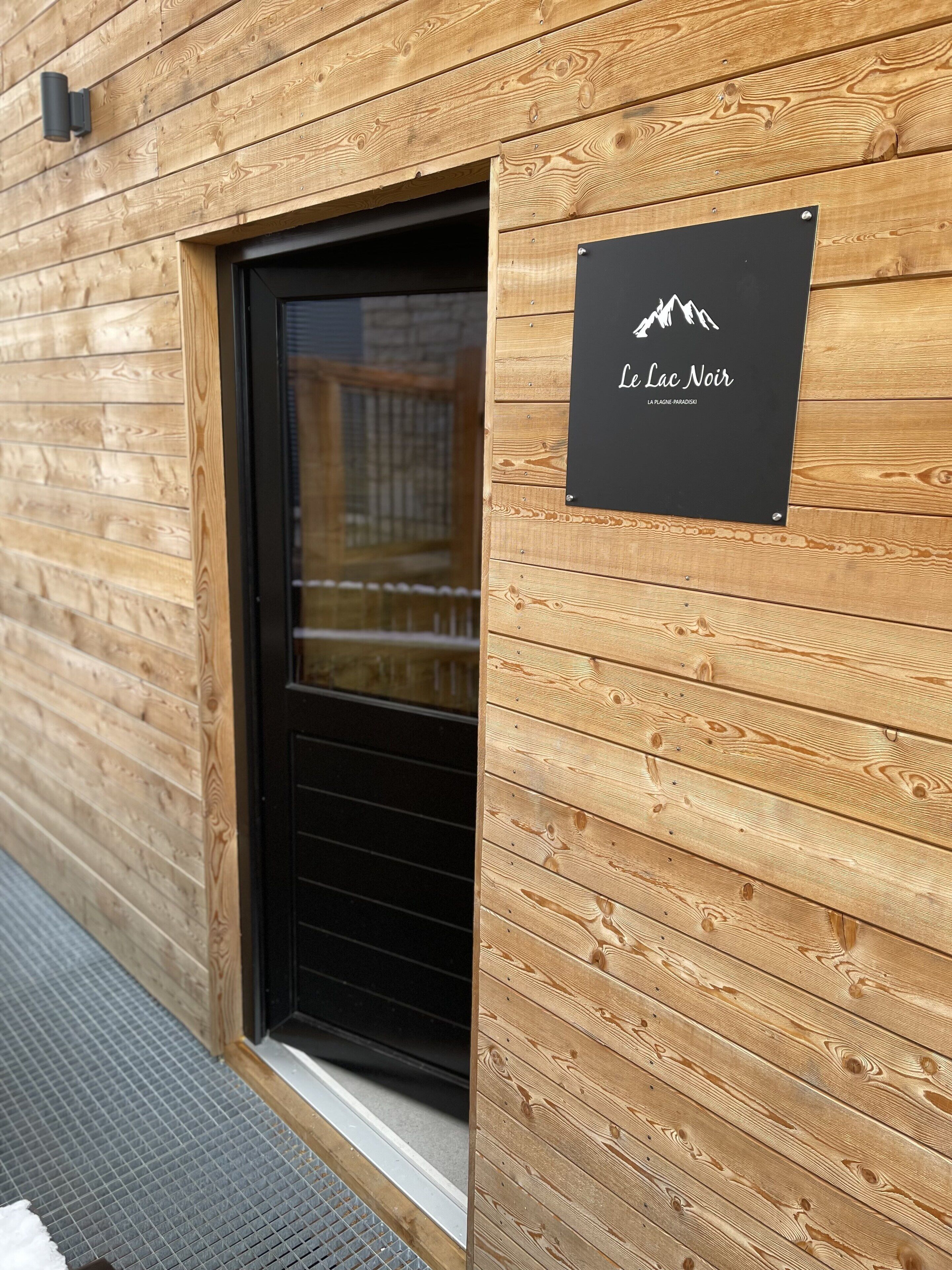 Un mur extérieur en bois, avec une porte noire et une enseigne sur laquelle on peut lire « Le Lac Noir ».