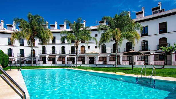 Una piscina con agua azul cristalina, rodeada de edificios blancos con balcones y palmeras.
