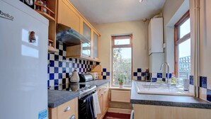 Een kleine keuken met een koelkast, fornuis en wastafel. Het heeft een betegelde spatwand met ruitpatroon, houten kastjes en een raam dat voor natuurlijk licht zorgt.