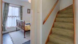 Een trap met beklede treden en houten leuningen, die leidt naar een kamer met een bed, een raam met gordijnen en een wandlamp.