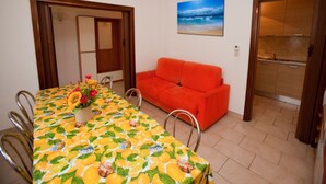 A dining area with a table covered in a fruit pattern, a red sofa, and a painting of a beach scene. - Spacious flat 50m from the beach (Rosolina Mare)