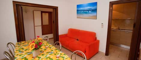 A dining area with a table covered in a fruit pattern, a red sofa, and a painting of a beach scene.