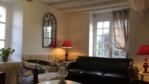 A cozy living room with a black leather sofa, a round white table with a candle, and two red lamps. A stone wall and a window with curtains are visible.