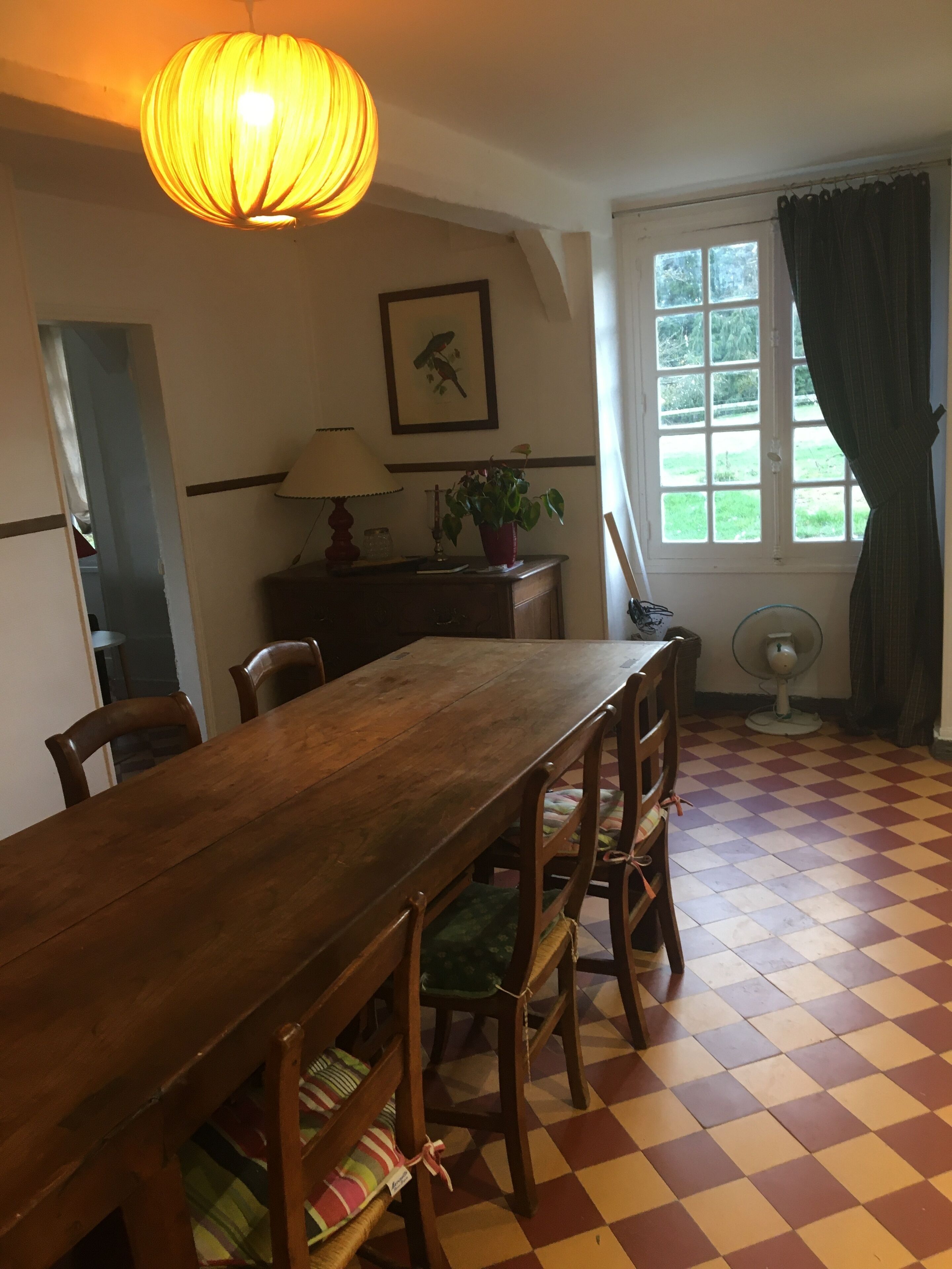 Un comedor con una mesa y sillas de madera, una lámpara, una planta en maceta, una ventana con cortinas y un ventilador.