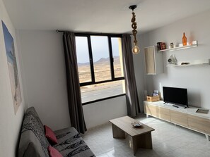 Ein Wohnzimmer mit einer Couch, einem Couchtisch, einem Fernseher und einem Fenster mit Blick auf eine Straße und Berge.