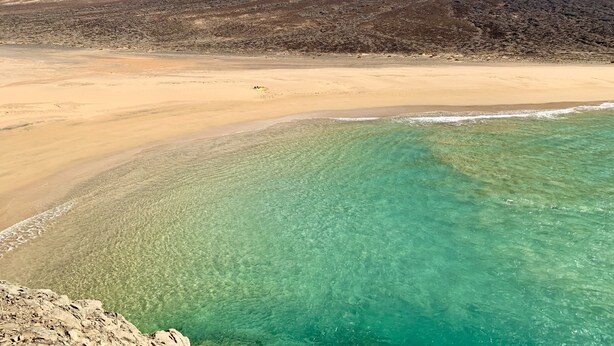 En strand med kristallklart turkost vatten, en sandstrand och ett bergigt landskap i bakgrunden.