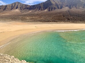 Ein Strand mit klarem türkisblauem Wasser, einem Sandufer und einem bergigen Hintergrund.
