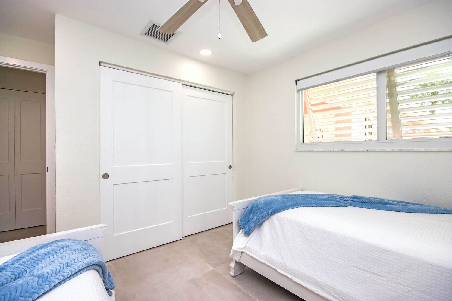 A bedroom with two beds, a window with blinds, a ceiling fan, and a white door.