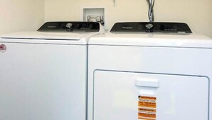Un espace buanderie comprenant deux machines à laver à chargement frontal de couleur blanche, une étagère avec une machine à café et une multiprise dotée de plusieurs câbles.