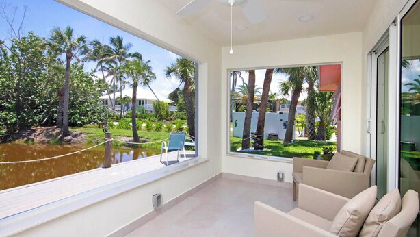 Een ruim balkon met uitzicht op palmbomen en een waterpartij, een plafondventilator en een beige bank.