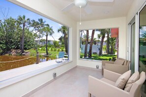 A spacious balcony with a view of palm trees and a body of water, a ceiling fan, and a beige sofa.