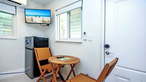 Una habitación compacta con refrigerador, televisor montado en la pared, una pequeña mesa redonda y dos sillas de mimbre, además de una puerta con ventana.