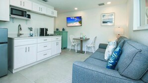 A compact living space with a kitchenette, a grey sofa with a blue pillow, and a dining area with a table and chairs. - West End Paradise Unit Coral 4 - One bedroom efficiency suite (Sanibel)