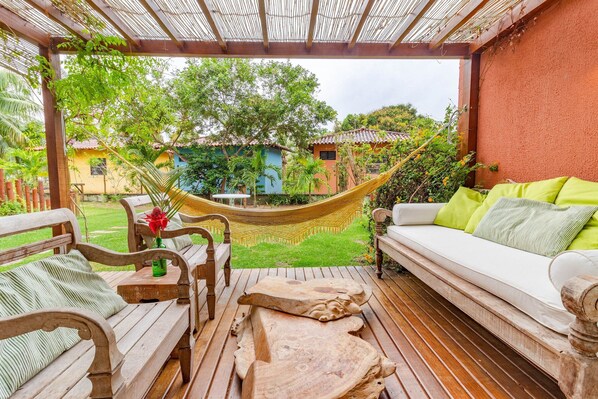A wooden deck with a hammock, a white sofa with green cushions, and a wooden table with a vase of flowers. - MSP01 Apt 3 Quartos Pé na Areia (Cairu)